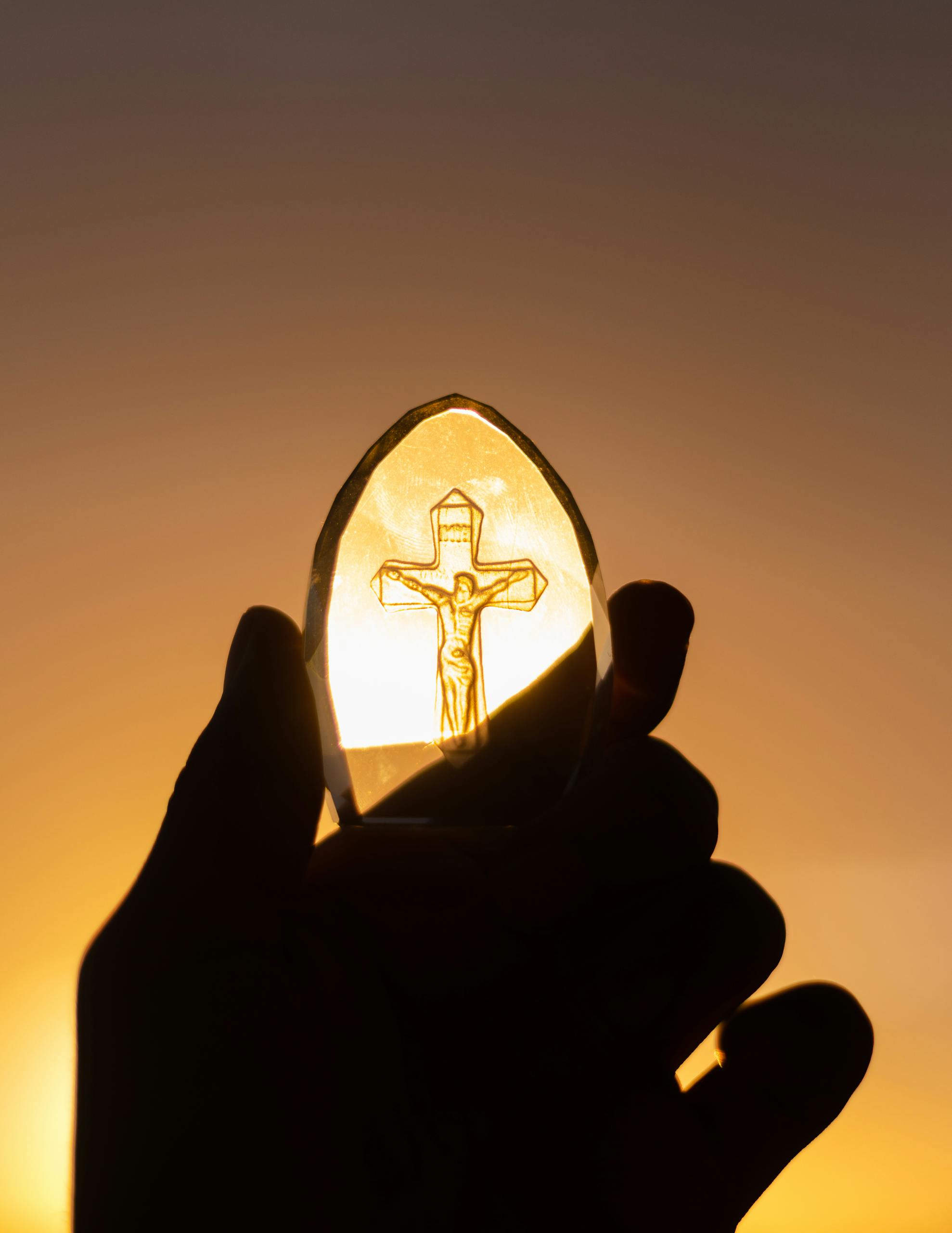 A silhouetted hand holds a crucifix against a vibrant sunset sky, symbolizing faith and spirituality.
